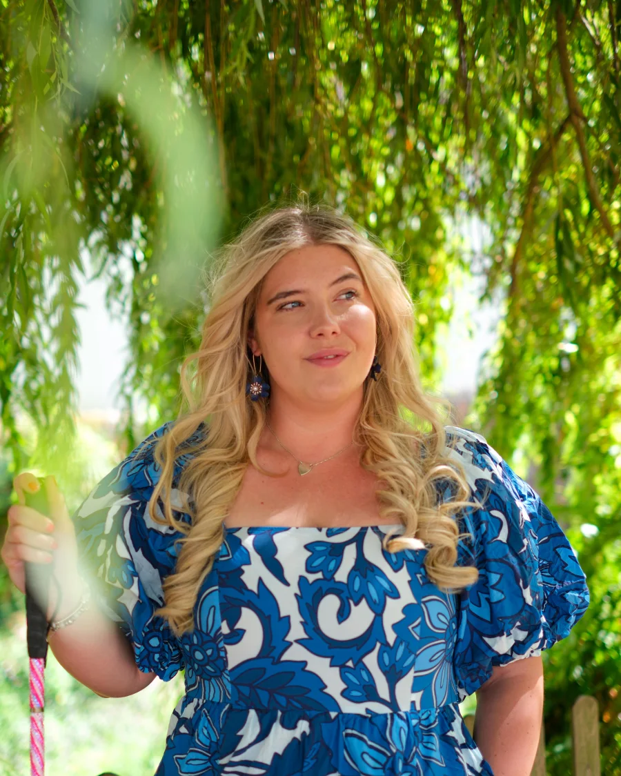 Tilly Dowler in a blue and white floral dress under a willow tree, holding her cane.