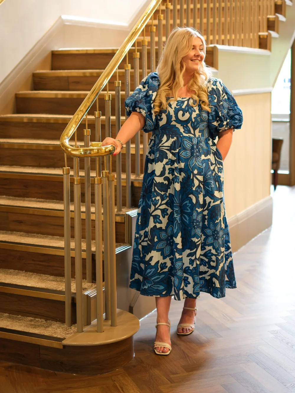 Tilly Dowler in a blue floral midi dress on a staircase, one hand on the brass handrail.